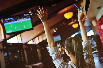 Foto op Canvas young pretty girl football fan with a glass of beer in hands watching football in the sports bar © Evgeniy Kalinovskiy