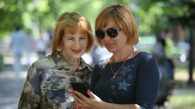 Portrait of a wise mother and her fashionable blond woman in sunglasses looking through  photos in their mobile in a green alley in summer 