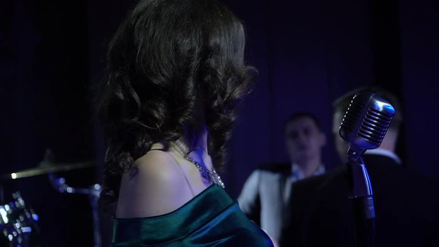 A beautiful girl stands on stage sings in a microphone and dance in blue ligts on the background of the jazz band