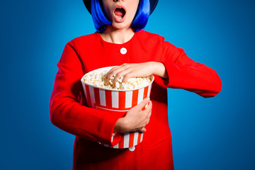 Naklejka premium Cropped portrait of shocked scared girl in red coat with wide open mouth having problem trouble panic holding big bucket of pop corn in hands looking spectacular program isolated on blue background