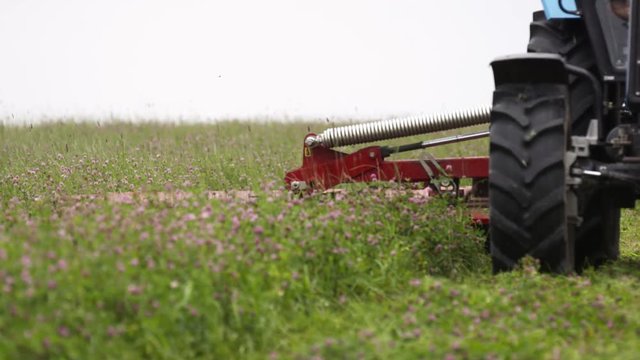 Tractor riding on farm field with agriculture combine attachment cutting grass on cloudy summer day