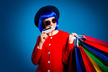 Portrait of pretty girl holding many colorful packets in hand biting plastic bank card with teeth isolated on blue background with shadow and light, comfortable quickly shopping concept