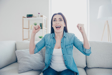 Portrait of impressed glad woman in denim outfit holding hands up yelling celebrating victory sitting on sofa in modern white living room