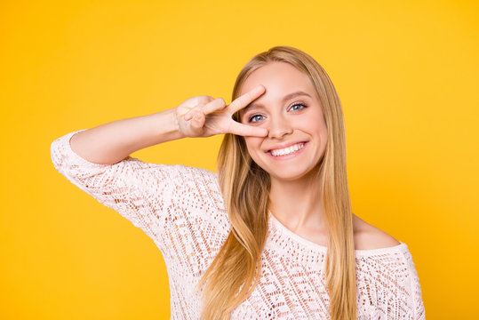 Portrait of cheerful toothy girl with white smile gesturing v-sign peace symbol near eye looking at camera isolated on yellow background