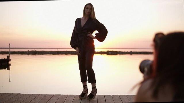 Beautiful tall model posing for the camera in black rompers and sandals standing in a full length in front the sea. Photoshooting concept