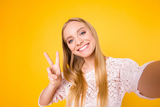 Self portrait of charming cheerful girl shooting selfie on front camera gesturing v-sign peace symbol with fingers isolated on yellow background