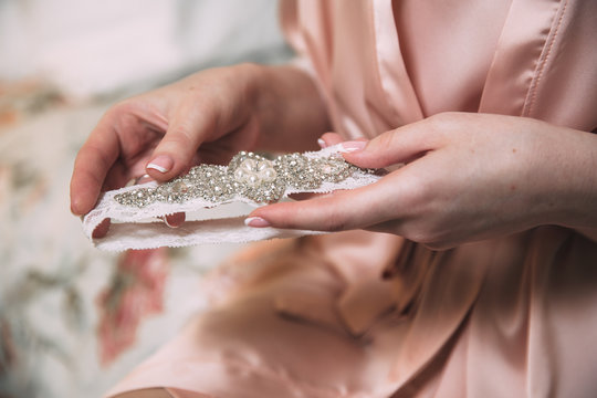Morning of the bride. Close-up of young bride in pink dressing-gown putting on white garter at the wedding day. Hands of the girl with accessories.