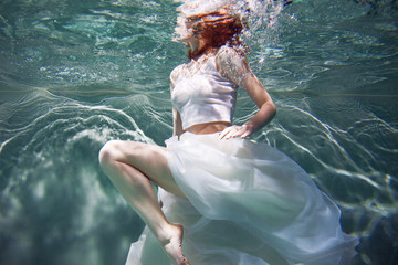 Underwater girl. Beautiful red-haired woman in a white dress, swimming under water.