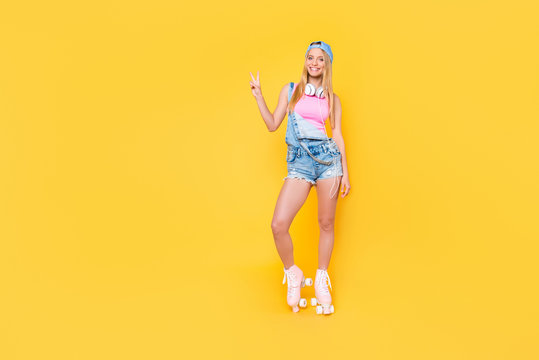 Full size portrait of positive funky girl in baseball cap denim overall with headset on neck on roller skates gesturing v-sign with hand looking at camera isolated on yellow background