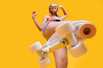 Bottom view of roller skate step on camera, cheerful joyful playful funky girl showing equipment for fitness workout isolated on yellow background - obrazy, fototapety, plakaty