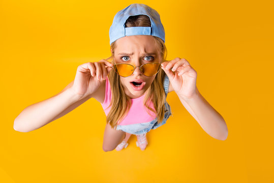 WTF! Top view portrait of sad disappointed girl looking out glasses on face holding eyelets with two hands isolated on yellow background