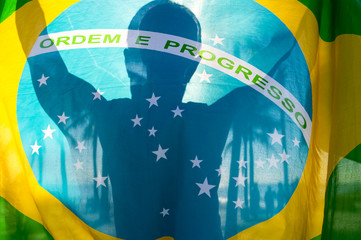 Backlit shadow silhouette of man holding Brazilian flag in bright sunlight with palm trees. 