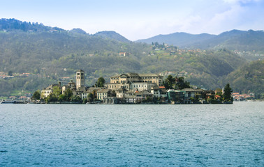 Fototapeta premium Island with the large villa, church and residential buildings in the sea. In the foreground is blue sea water, the focus is the island, in the background are mountains with forest and homes in between