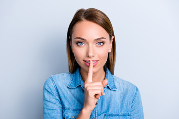 Shh! Portrait of mysterious cute girl in jeans shirt holding forefinger on her lips asking to keep silence isolated on grey background
