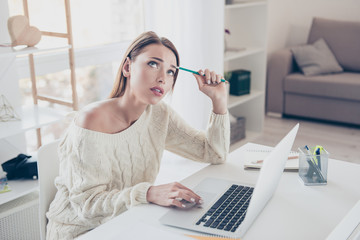 Portrait of thoughtful dreamy woman looking up trying to find solution decision holding pencil in hand browsing on laptop sitting in modern home-office with interior