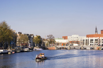 View on river Amstel in Amsterdam, capital of the Netherlands, with a boat, blue sky, trees and buildings