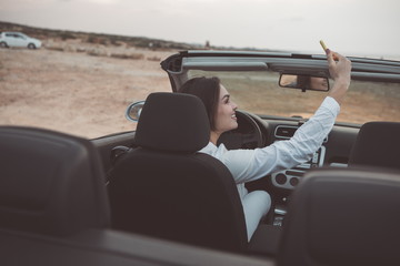 Cheerful young woman is making selfie during her summer trip. She is sitting at steering wheel in her vehicle and smiling. Lady is looking at cellphone camera with joy 