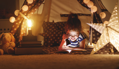 happy child girl laughing and reading book in dark in tent at home