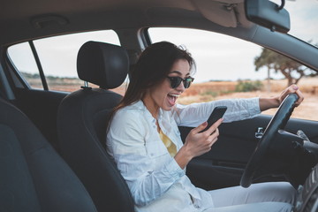 Excited young woman is reading message on smartphone and laughing. She is sitting at steering wheel of her car with relaxation. Beautiful natural landscape is on background 