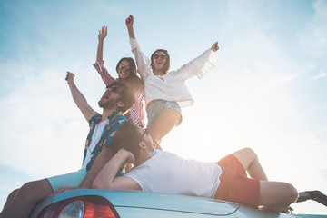 Lets enjoying life together. Low angle of happy friends enjoying summer vacation. Man is lying on car with relaxation. Girls are standing on transport and dancing while smiling 