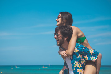 Fototapeta na wymiar Side view of excited loving couple is enjoying summer vacation together. Man is holding woman on his back and smiling. They are looking at the sea with enjoyment 