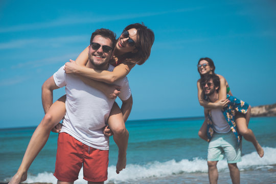 This is our best vacation. Portrait of happy young man is carrying his girlfriend on back and laughing. Their friends are having fun on background near the sea 