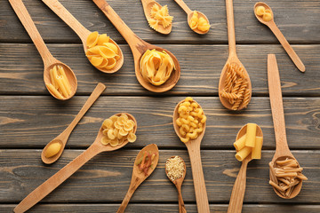 Spoons with different uncooked pasta on wooden background