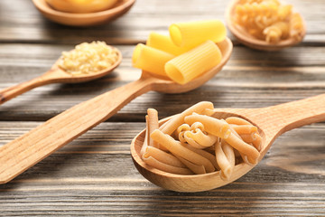 Spoons with different uncooked pasta on wooden background