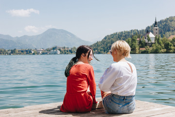 Fototapeta na wymiar back view of stylish girls sitting on wooden pier near tranquil mountain lake, bled, slovenia