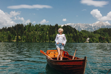 Fototapeta na wymiar beautiful young woman in sunglasses standing in boat at tranquil mountain lake, bled, slovenia