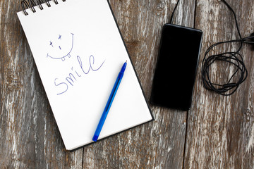 White notebook with a blue pen is lying on the old vintage background. Smile emotions is drawn and written on sketch book. Black smartphone with earphones is lying nearby. Modern technologies.