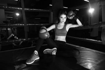 Full body portrait of boxer woman with long dark hair pulled back in pony tail, wearing pink sports bra, leggings, trainers and red boxing gloves, sitting in corner of ring and leaning on ropes.
