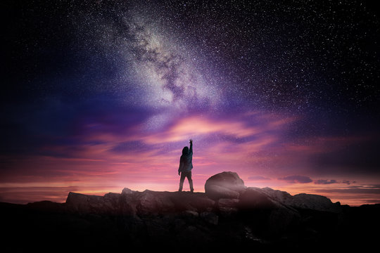Night time long exposure landscape photography. A man standing in a high place reaching up in wonder to the Milky Way galaxy, photo composite.