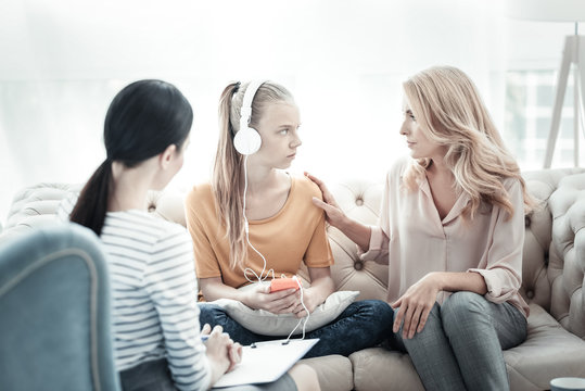 Problems with communication. Pleasant worried mother sitting with female psychologist and teen girl listening to music