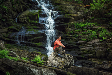 Tourist sits on a rock near a forest waterfall.