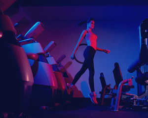 Young slim brunette woman dressed in sports apparel jumping among rows of exercise machines. Female model wearing leggings, top and trainers illuminated by pink and purple light posing at gym.