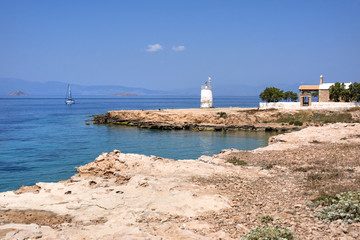 Fototapeta na wymiar Greece, Aegina: Panoramic view of calm beautiful coastline of the famous Greek island with clear ocean water, sailing boat, white lighthouse, horizon and small chapel and blue sky in the background.