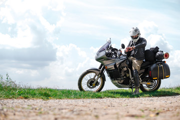 Rider Man looks at a watch and off road adventure motorcycles with side bags and equipment for long road trip, river and clouds on background, enduro travel touring concept