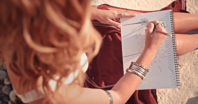 Hippie woman with silver jewelry drawing in sketchbook at beach