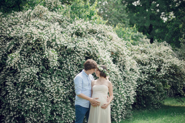 Happy pregnant woman and her husband hugging and kissing near the white flower bush in the green park on sunny summer day. Happy family