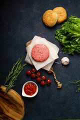 Cooking burger: classical american burger ingredients on dark blue background with copy space. Raw beef patties, vegetables, buns, lettuce.