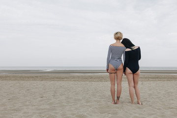 rear view of attractive young women in bodysuits embracing on sandy beach on cloudy day