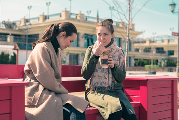 Two young women sit on a bright pink bench and drink coffee in cups, enjoying the Sunny weather and foam from coffee