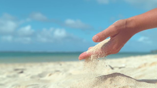 SLOW MOTION, CLOSE UP: Tiny grains of white sand get swept away from female hand by the gentle summer breeze. Unrecognizable woman on vacation playing by the ocean scoops up a handful of hot sand.