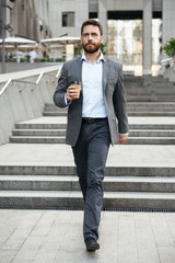 Photo of successful businessman 40s in gray suit walking down stairs of modern business center with takeaway coffee in hand