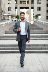 Photo of pleased entrepreneur man 40s in gray suit walking down stairs of modern business center, with takeaway coffee in hand