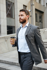 Photo closeup of successful entrepreneur or director man 40s in gray suit holding takeaway coffee in hand, and walking down stairs of modern business center