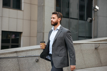 Profile photo of successful executive director or businessman 40s in gray suit holding takeaway coffee, and walking along street with modern business center background