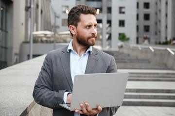 Image of caucasian office man 40s in classical suit holding open silver laptop, while standing in front of business center in big city