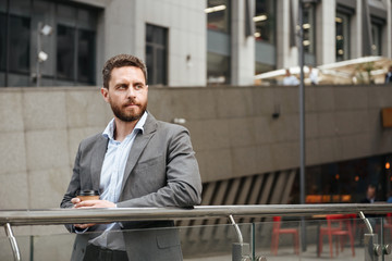 Image of successful entrepreneur man 40s in formal wear looking aside, while standing and drinking takeaway coffee in front of modern business center